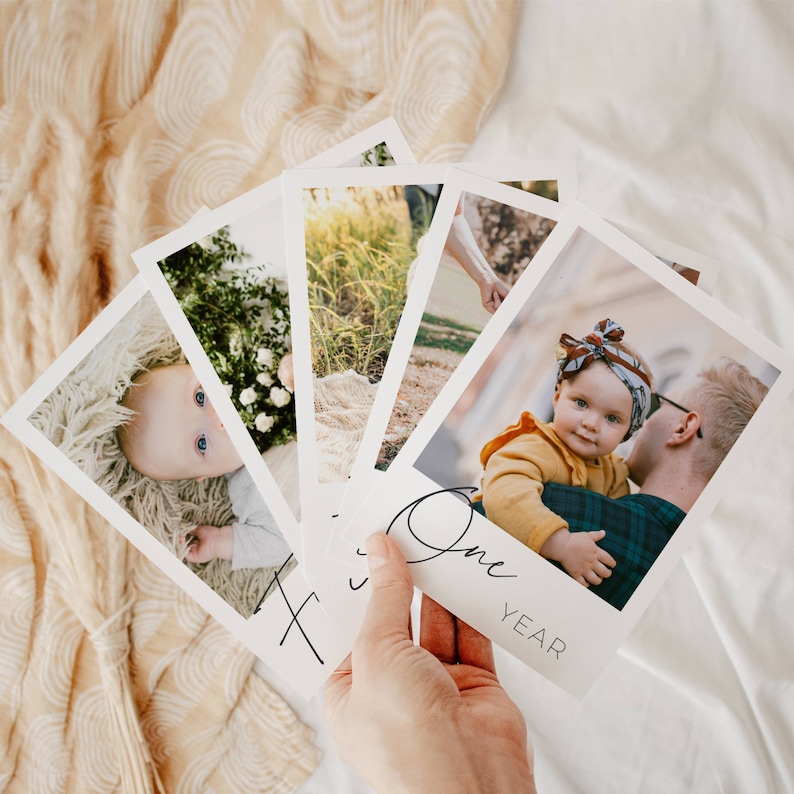 May include: A set of five white photo cards with black text and images of a baby. The cards feature photos of the baby in different settings, including a field of grass, a park, and a close-up of the baby's face. The text on the cards reads "One Year".
