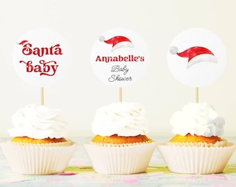 Könnte beinhalten: Drei Cupcakes mit weißem Zuckerguss und dekorativen Toppern. Einer der Topper trägt die Aufschrift "Santa baby" in roter Schrift, ein anderer "Annabelle's Baby Shower", und der dritte zeigt eine Illustration eines Weihnachtsmützen. Die Cupcakes sind in weißen Papierförmchen.