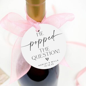 May include: White circle tag with black text that reads "He popped the question!" with a small heart symbol and the text "Harley & Willa 10.1.25" below. The tag is tied to a bottle of wine with a pink ribbon.
