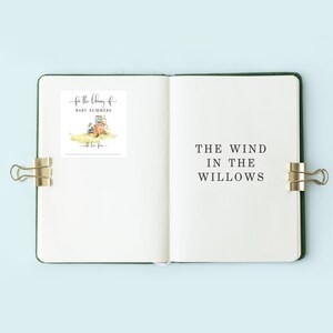 May include: Open journal with the title "The Wind in the Willows" printed on the right page. A sticker on the left page reads "For the library of Baby Summers" with a watercolor illustration of books and a tree stump. Gold binder clips secure the pages.