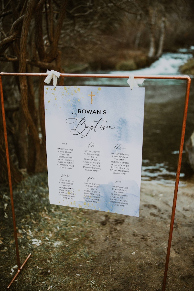 May include: A baptism seating chart with a watercolor design in blue and gold. The sign is held by a copper-colored metal stand. The text reads "Rowan's Baptism" with guest names listed below.