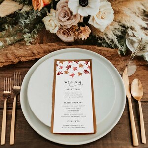 May include: A menu card with a brown border and a white interior, featuring a fall leaves design. The menu lists appetizers, main courses, and desserts. The card is placed on a white plate with gold silverware and a floral arrangement in the background.