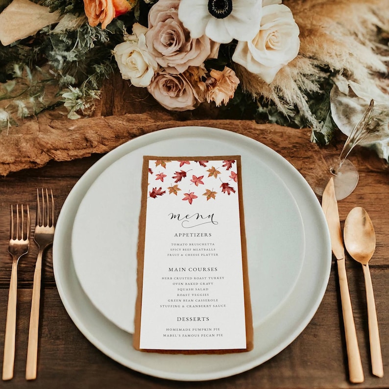 May include: A menu card with a brown border and a white interior, featuring a fall leaves design. The menu lists appetizers, main courses, and desserts. The card is placed on a white plate with gold silverware and a floral arrangement in the background.