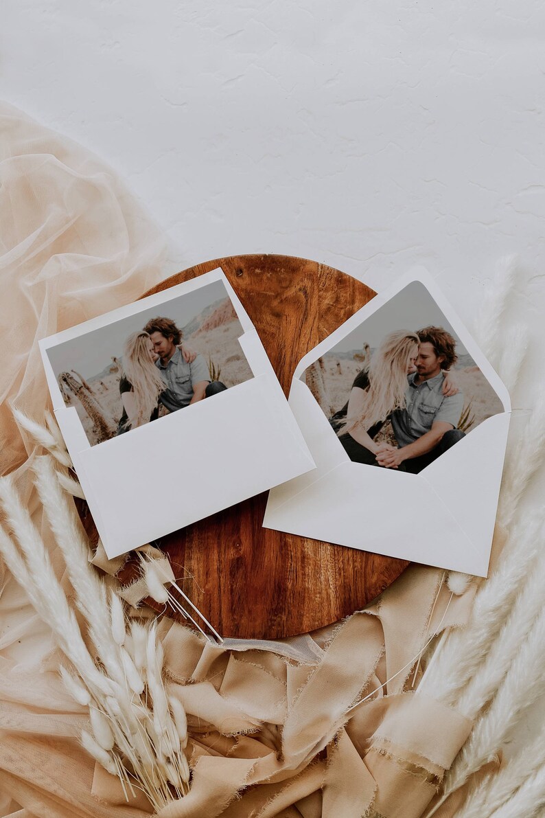 May include: Two white envelopes with a photo of a couple embracing on a desert landscape. The envelopes are on a wooden surface with a beige fabric and white dried flowers.