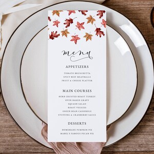 May include: A white menu card with a fall leaf design, featuring appetizers, main courses, and desserts. The menu is set on a white plate with a wooden background. The menu includes items like roast turkey and pumpkin pie.