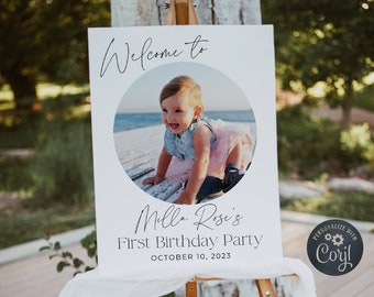 May include: White welcome sign with a photo of a baby girl in a blue and pink dress. The sign reads "Welcome to Milla Rose's First Birthday Party October 10, 2023".
