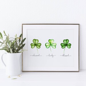 May include: A framed print with three watercolor shamrocks. The shamrocks are green and have a white background. The words "charmed", "lucky", and "blessed" are written below the shamrocks.