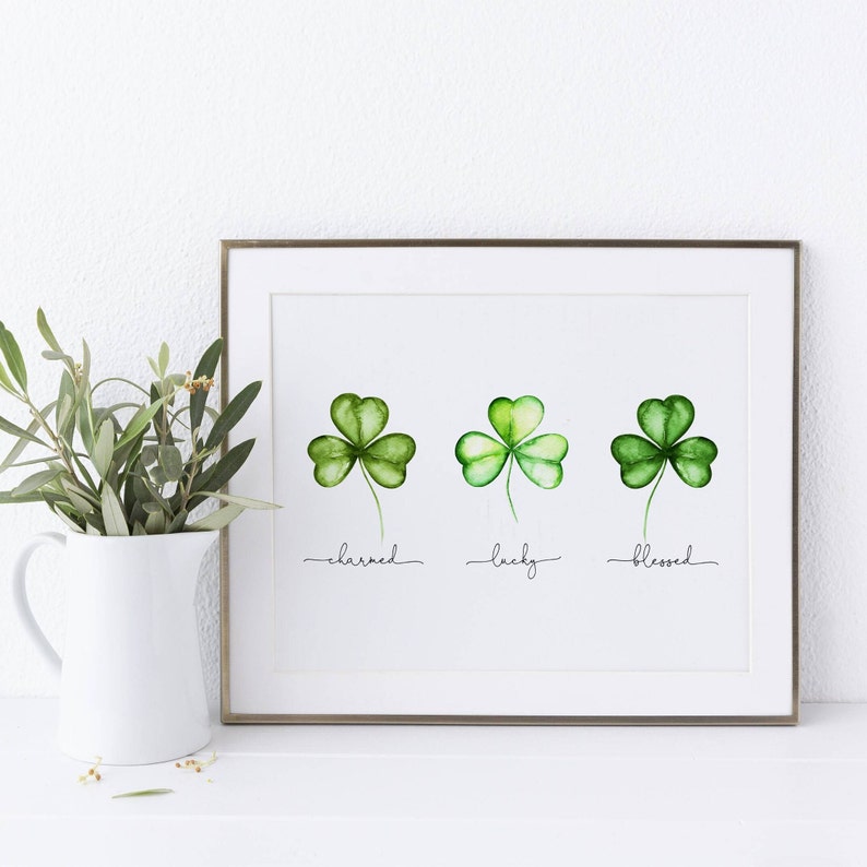 May include: A framed print with three watercolor shamrocks. The shamrocks are green and have a white background. The words "charmed", "lucky", and "blessed" are written below the shamrocks.