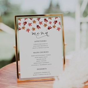 May include: A gold-framed menu card with a white background, featuring a fall leaf design at the top. The menu lists appetizers, main courses, and desserts, including pumpkin pie. The menu is displayed on a wooden surface.