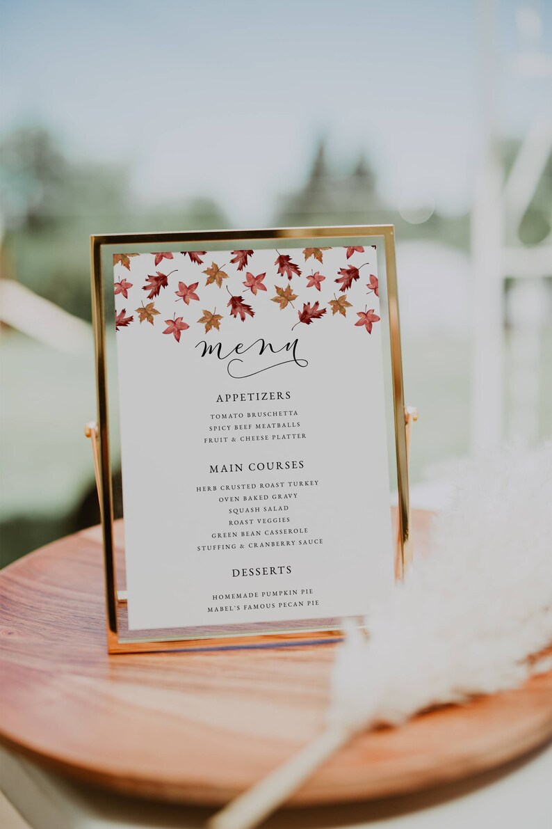 May include: A gold-framed menu card with a white background, featuring a fall leaf design at the top. The menu lists appetizers, main courses, and desserts, including pumpkin pie. The menu is displayed on a wooden surface.
