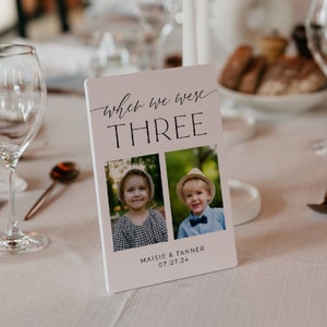 May include: A white table card with the text "when we were three" in black script font. Two photos of children are below the text, with the names "Maisie & Tanner" and the date "07.07.24" below the photos.