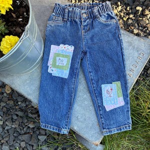 May include: Medium-wash blue denim jeans featuring an elasticated waistband and two decorative fabric patches. The patches are composed of layered floral and solid-coloured fabric squares, each displaying a small embroidered chicken motif.