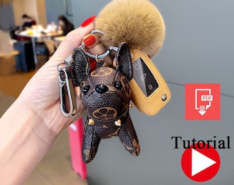 Op de afbeelding: Een bruine en zwarte kunstleren Franse bulldog sleutelhanger met een gouden ketting en een bruine bontpompon. De sleutelhanger bevat ook een bruine leren sleutelhouder met een zilveren knop.