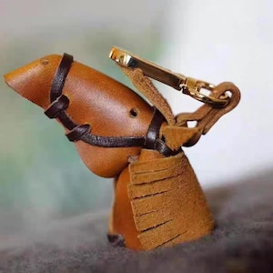 Puede incluir: Un llavero de cuero marrón en forma de cabeza de caballo con un anillo de metal dorado y una franja de cuero marrón.