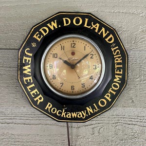 May include: Black octagonal clock with gold lettering that reads "EDW. DOLAND JEWELER OPTOMETRIST Rockaway, N.J." The clock face is beige with black numerals and hands. A brown cord hangs below the clock.