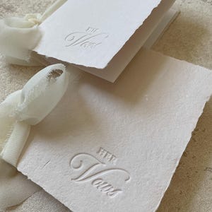 May include: Close-up of a stack of white paper cards with the embossed text "HER Vows". The cards have a deckled edge and are tied with a cream-colored ribbon. The paper has a textured, handmade appearance.