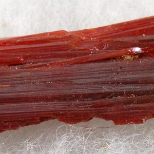 Könnte beinhalten: Nahaufnahme einer leuchtend roten, durchscheinenden Mineralprobe. Die Kristallstruktur besteht aus langen, parallelen, nadelförmigen Formationen. Die Farbe reicht von einem tiefen, satten Rot bis zu einem etwas helleren Farbton mit glänzendem Aussehen. Das Mineral liegt auf einer weißen, faserigen Oberfläche.