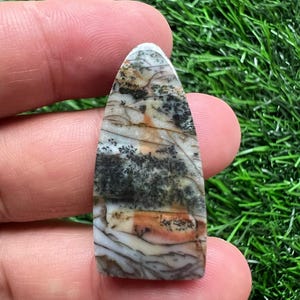 Puede incluir: Una gema pulida y alargada con una mezcla de colores. La piedra presenta un patrón de tonos negros, blancos y anaranjados, con un acabado liso y brillante. La forma es un rectángulo redondeado, ideal para joyería o exhibición.