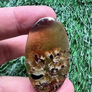 Peut inclure: Une pierre précieuse ovale avec un mélange de couleurs marron, vert et crème. La pierre a une qualité translucide avec des motifs et des inclusions uniques. La pierre est tenue dans une main sur un fond d'herbe artificielle verte.