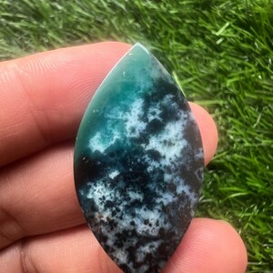 Puede incluir: Una piedra preciosa pulida con forma de lágrima. La piedra presenta una mezcla de colores, incluyendo verde azulado, negro y blanco, con un patrón moteado. La piedra preciosa se sostiene en una mano sobre un fondo de hierba verde.
