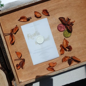 May include: A white wedding invitation with gold calligraphy that reads "Rosella & ..." and a white wax seal with a floral design. The invitation is on a wooden surface with dried orange flower petals.