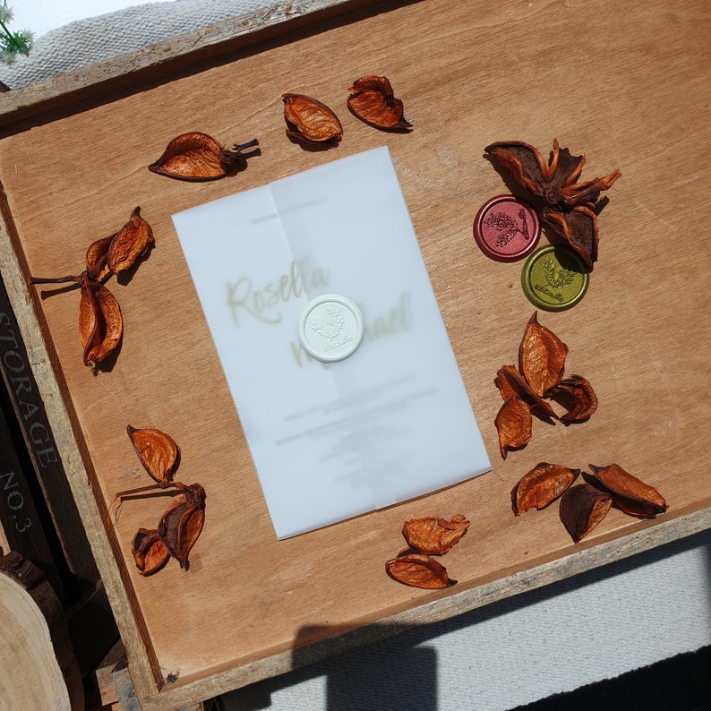 May include: A white wedding invitation with gold calligraphy that reads "Rosella & ..." and a white wax seal with a floral design. The invitation is on a wooden surface with dried orange flower petals.