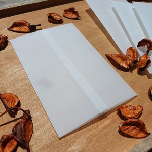 May include: A white card with a white border, folded in half. The card is lying on a wooden surface with dried leaves scattered around it.