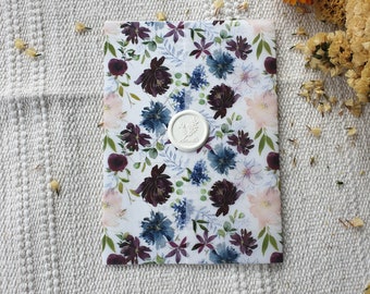 Puede incluir: Una tarjeta blanca con un patrón floral en tonos azules, morados y rosas. Un sello de cera blanco con un diseño está en el centro de la tarjeta.