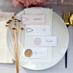 Puede incluir: Un plato blanco con cuatro tarjetas de lugar sobre un mantel de lino beige. Las tarjetas de lugar están decoradas con sellos de cera y caligrafía. Los nombres en las tarjetas de lugar son "Andreas", "Temi", "Elizabeth" y "Suzanne".