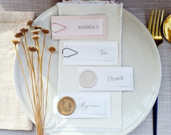 Op de afbeelding: Een wit bord met vier plaatskaarten op een beige linnen tafelkleed. De plaatskaarten zijn versierd met waszegels en kalligrafie. De namen op de plaatskaarten zijn "Andreas", "Temi", "Elizabeth" en "Suzanne".