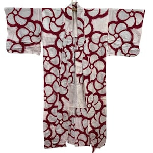 Puede incluir: Un kimono blanco con un patrón repetido de diseños florales granate. El kimono está colgado de una percha de madera sobre un fondo blanco. Las mangas son anchas y la prenda está abierta por delante.