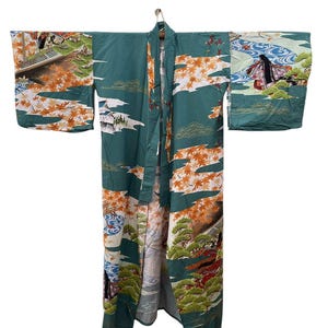 May include: A vintage teal kimono with a landscape design featuring orange autumn leaves, buildings, and figures. The kimono is hanging on a wooden hanger, showcasing its full length and intricate pattern. The sleeves and body are adorned with detailed artwork.