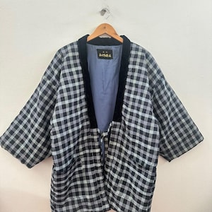 Könnte beinhalten: Eine gesteppte Jacke mit schwarz-weißem Karomuster, weiten Dreiviertelärmeln und einem schwarzen Samtbesatz entlang der Vorderseite. Das Innere ist mit hellblauem Stoff gefüttert und enthält ein schwarzes Etikett mit goldfarbenem Text.