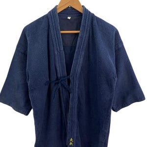 May include: A navy blue martial arts uniform jacket, hanging on a wooden hanger. The jacket has a textured weave, short sleeves, and a tie closure. The Japanese characters are embroidered in gold on the front.