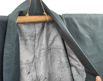 May include: A dark teal kimono with a geometric pattern. The interior lining features a gray and white illustration of a bridge scene with figures. The kimono is hanging from a wooden rod with a twine hanger.