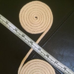 May include: Two beige rope coils connected by a length of rope, with a measuring tape laid across them. The tape shows measurements in inches, with the rope coils appearing to be approximately 25 cm in diameter.