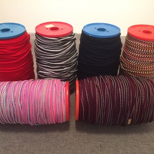 May include: Six spools of colourful cord, each wound around a red plastic spool with a blue plastic top. The cords are red, black and white striped, black, gold and white striped, pink and white striped, and burgundy and white striped.