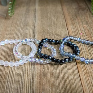 Pode incluir: Quatro pulseiras de contas, duas brancas, uma iridescente e uma preta com detalhes iridescentes. As pulseiras estão empilhadas sobre uma superfície de madeira.