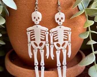 May include: A pair of white skeleton earrings with silver hooks. The skeletons are dangling and have a glittery finish.