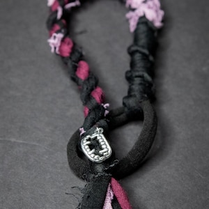 May include: A black and purple braided necklace with a silver charm in the shape of a mouth with teeth. The necklace is made of fabric and has a frayed edge.