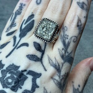 May include: Silver ring with a rectangular pyrite stone. The pyrite is a mosaic of small, reflective pieces. The ring has a beaded border and a wide band. The ring is worn on a finger with black floral tattoos.