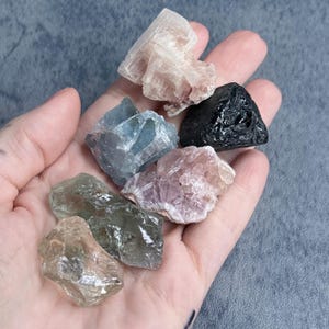 May include: A collection of raw crystals in various colors, including pink, blue, black, and clear. The crystals are held in a hand, showcasing their natural textures and shapes. The crystals are a mix of sizes and shapes, with a focus on their natural beauty.
