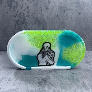 May include: A white oval trinket dish with a teal and green swirl design and a black and white sticker of a skeleton hugging a person. The dish is covered in green glitter.