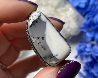May include: A silver ring with a white and gray dendritic opal stone. The stone has a unique pattern of black dendrites.