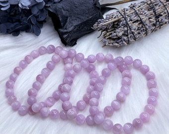 Puede incluir: Cuatro pulseras de piedras preciosas de color lavanda hechas de cuentas redondas. Las pulseras están apiladas una encima de la otra.