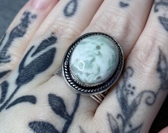 May include: A silver ring with an oval, light green and white stone. The stone is set in a twisted rope-like border. The ring is on a hand with tattoos.