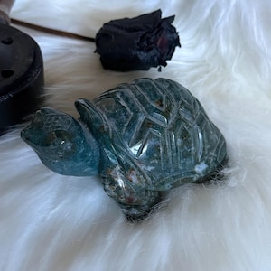Peut inclure: Une figurine de tortue en pierre sculptée dans les tons de vert et de bleu. La tortue présente des motifs de carapace détaillés et une finition lisse et polie. La figurine est exposée sur une surface blanche et duveteuse.