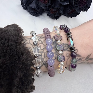 May include: Five bracelets stacked on a wrist. The bracelets are made of different materials, including lava rock, glass beads, and metal. The bracelets are in shades of purple, gray, and white.