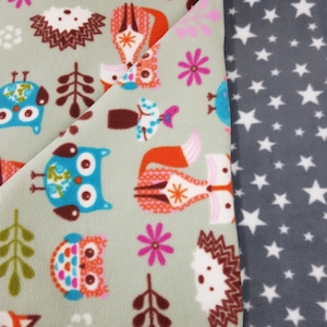 Può includere: Due coperte in pile. Una è verde chiaro con gufi, volpi, ricci, fiori e alberi a cartoni animati colorati. L'altra è grigio scuro con stelle bianche. Le coperte sono piegate l'una sull'altra.