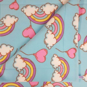 May include: A light blue fleece blanket featuring a repeating pattern of rainbows, clouds, and heart-shaped balloons. The rainbows have yellow, orange, and purple arches. The clouds are white, and the balloons are pink.